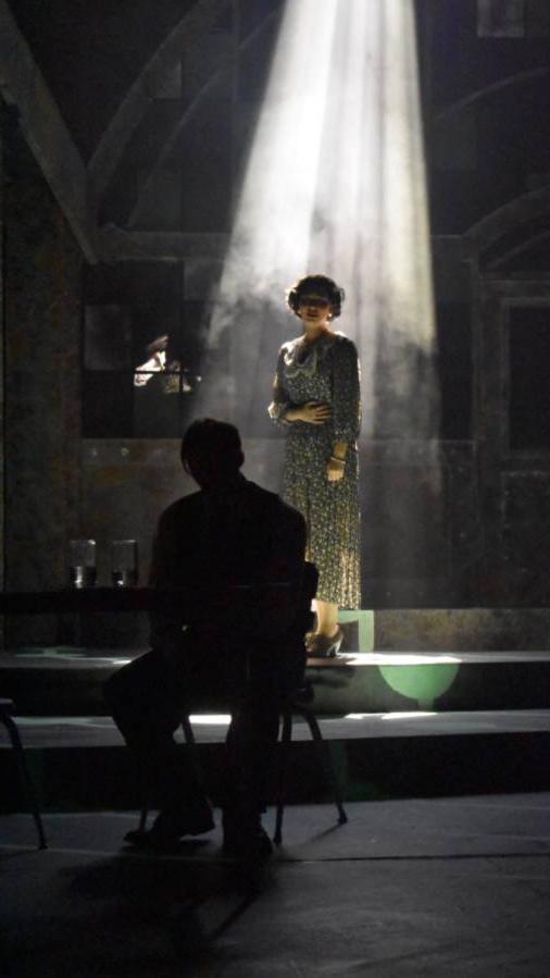 Image of lady standing in dark room with a ray of sunlight shining down on her while the sillouett of a man is sitting in front of her facing away, head down